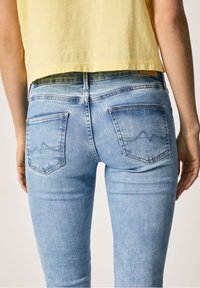 Jeans de mezclilla azul claro con corte ajustado, con dos bolsillos traseros y un sutil patrón de montañas en el bolsillo derecho.
