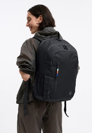 Frau lächelnd, trägt eine dunkle Jacke und Hose, trägt einen großen schwarzen Rucksack mit Frontreißverschluss und seitlichen Netzfächern.