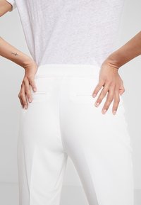Witte op maat gemaakte broek met een zachte textuur, voorzien van twee achterzakken en een aansluitende tailleband. Het shirt is licht en licht doorschijnend.