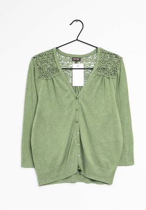 Cardigan verde con bottoni, maniche lunghe e dettagli di pizzo floreale sulle spalle e sulla parte superiore della schiena, appeso a una gruccia nera.