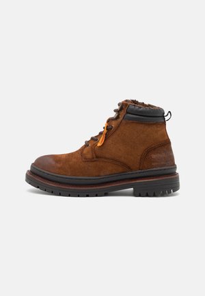 Bottines en suède marron avec un col en cuir noir, dotées de lacets orange et d'une semelle en caoutchouc texturée avec des crampons profonds pour une traction optimale.