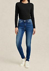 Mujer vestida con jeans skinny de tiro alto azules, camiseta negra de manga larga y zapatillas blancas de pie frente a un fondo liso.