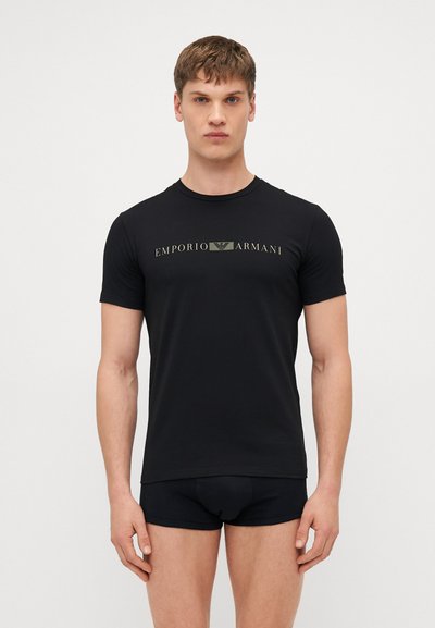 Crna pamučna majica s kratkim rukavima, okruglim izrezom, s centralnim zlatnim logotipom Emporio Armani. Model nosi crna gaćice.