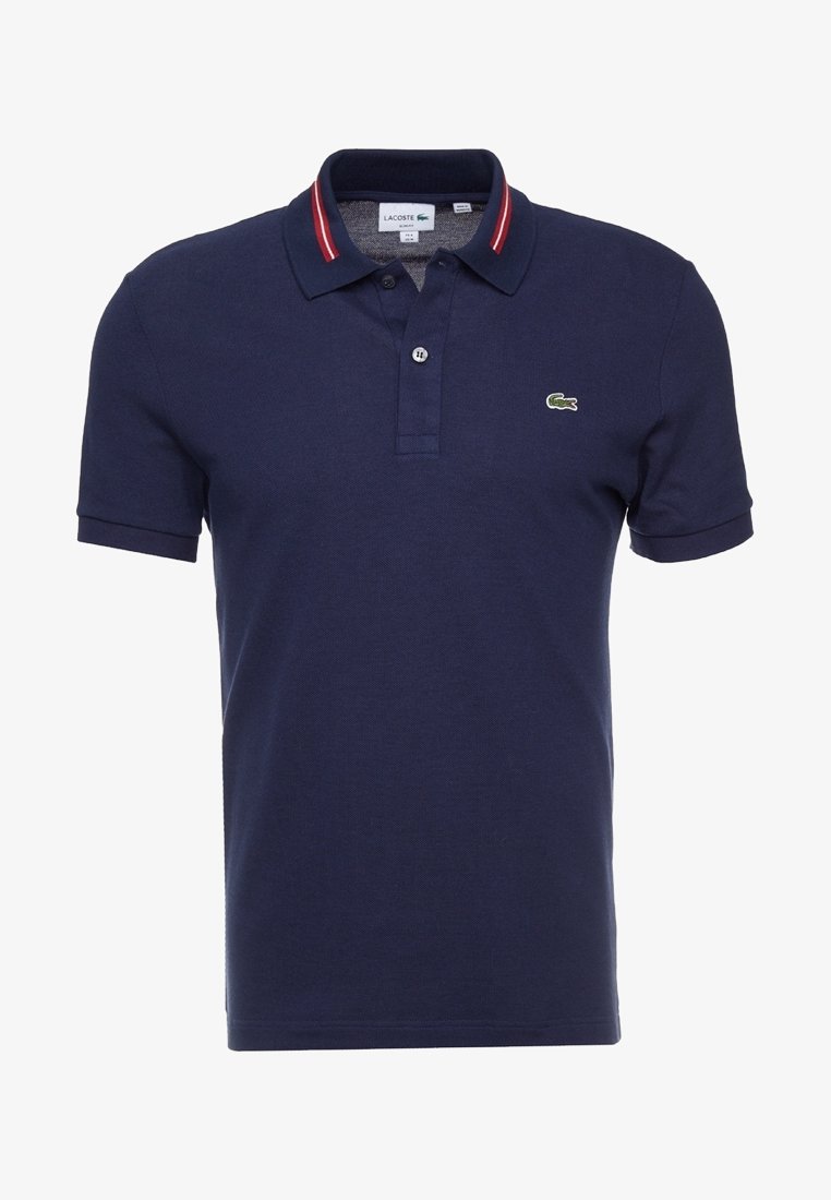 Polo Lacoste bleu marine avec col rayé rouge et blanc, patte de boutonnage à deux boutons, manches courtes, et petit logo crocodile vert sur la poitrine.