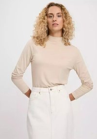 Beige long-sleeve turtleneck top with fitted silhouette, soft fabric, and ruched detailing on sleeves, paired with white high-waisted denim skirt.