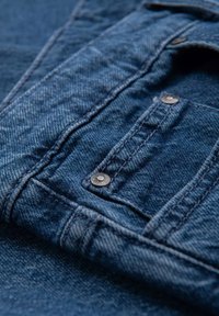 Primer plano de tela de mezclilla azul que muestra detalles de costura y remaches metálicos en un bolsillo.