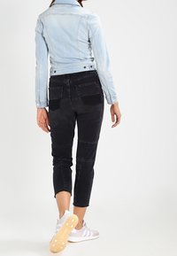 Hellblaue Jeansjacke, schwarze Cropped-Hose mit strukturierten Einsätzen und graue Turnschuhe mit weißen Streifen, von hinten gesehen.