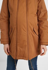 Veste matelassée marron avec deux poches à rabat à l'avant, doubles fermetures éclair en bas, portée sur un jean bleu par une personne debout, les mains détendues.
