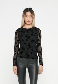 Sort langærmet top med blomster-velour mønster på kroppen og gennemsigtige meshærmer. Matchet med skinnende mørkegrønne tætsiddende bukser.