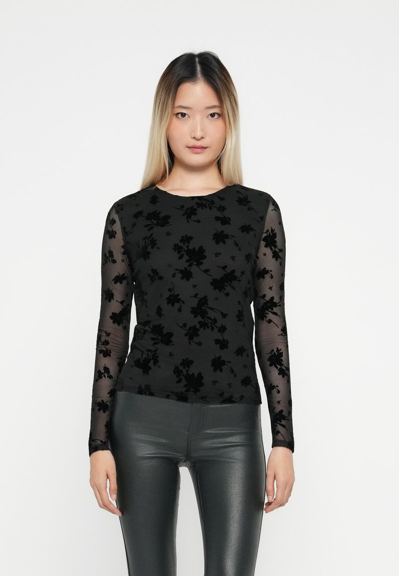 Sort langærmet top med blomster-velour mønster på kroppen og gennemsigtige meshærmer. Matchet med skinnende mørkegrønne tætsiddende bukser.