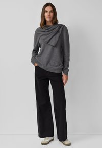 Grauer Pullover mit asymmetrischem, drapiertem Ausschnitt, kombiniert mit schwarzen Hosen mit weitem Bein und beigen Sneakers. Glatte Textur, lässiges Design.