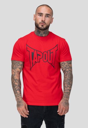 Homme tatoué portant un t-shirt rouge "Tapout" et un jean noir, debout devant un fond uni gris clair.
