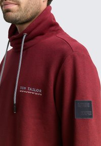 Rode sweatshirt met een hoge kraag, voorzien van contrasterende trekkoorden, een logo op de borst en een stoffen label op de mouw. Zachte textuur.