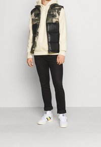 Vadderad svart väst med camouflage-design, gräddfärgad hoodie, svarta jeans och vita sneakers med gröna och gula ränder.