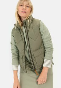 Olijfgroene puffer vest met een hoge kraag, rits aan de voorkant en twee voorzakken. Gelaagd over een mintgroene top met gestreepte manchetten. Gladde textuur.