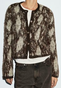 Kardigan mit braunem und cremefarbenem Muster, rundem Ausschnitt, vorderem Knopfverschluss und flauschiger Textur, kombiniert mit einem weißen Hemd und dunklen Jeans.