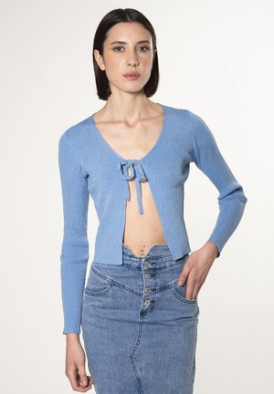 Hellblaues, geripptes Strickjacke mit V-Ausschnitt und vornem Schleifchen-Detail. Lange Ärmel und cropped Design, kombiniert mit einem Jeansrock mit Knöpfen.