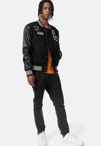 Svart varsityjacka med läderärmar, randiga manschetter och broderade applikationer, tillsammans med en orange t-shirt och svarta jeans. Vita sneakers.