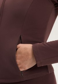 Barna sportos dzseki sima textúrával, cipzáras oldalsó zsebekkel és testhez álló ujjakkal. A tervezés sík varrásokat tartalmaz a kényelem érdekében.