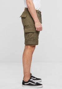 Alyvuogių žalios krovininės šortai su elastinga juosmens juosta, šoninėmis kišenėmis ir suktomis apačiomis, derinami su juodais sportbačiais su baltais akcentais.