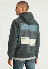 Felpa con cappuccio grigia. Presenta diverse toppe rettangolari in varie tonalità di blu e bianco con la scritta "CHASIN'" stampata.