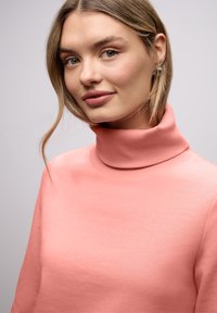 Pull à col roulé rose clair avec une texture lisse, col côtelé et manches longues. La coupe est décontractée et ample.