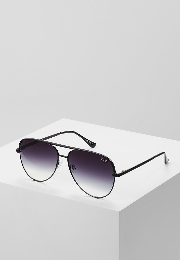 Des lunettes de soleil de style aviateur avec une monture en métal noir et des verres dégradés foncés, portant le nom de la marque "QUAY" affiché sur le verre.