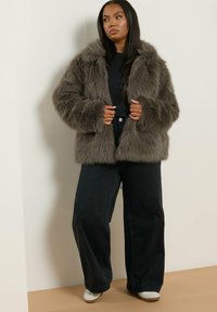 Giacca in finta pelliccia grigia con una consistenza morbida e cappuccio, caratterizzata da un fit rilassato, abbinata a pantaloni neri a gamba larga e scarpe bianche.