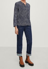 Persona che indossa una blusa a fantasia blu navy, jeans blu scuro con risvolti e stivaletti marroni con elastiche nere ai lati.