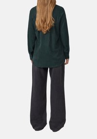 Vrouw met lang blond haar, gekleed in een donkergroene, getextureerde, lange mouw shirt en losse zwarte broek, staande met haar rug naar de camera tegen een effen achtergrond.