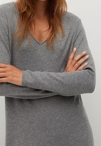 Femme avec de longs cheveux bruns portant un pull gris à manches longues et col en V, les bras croisés, fond neutre.
