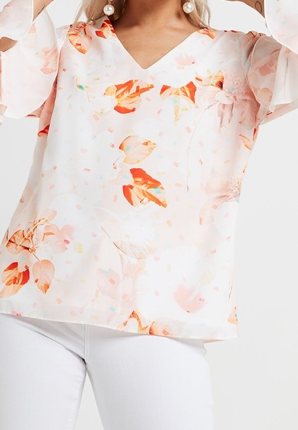 Femme portant un chemisier blanc avec des motifs floraux orange et rose et un pantalon blanc, posant avec les bras levés près de la tête.