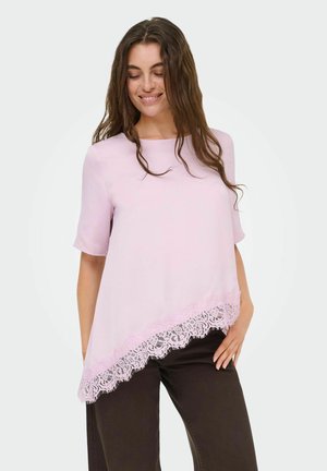 Žena s dlhými hnedými vlasmi, ktorá nosí svetloružový asymetrický top s čipkovou bordúrou a tmavé nohavice, usmievajúca sa so zavretými očami.