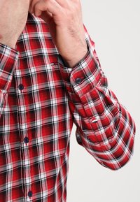 Mann in einem rot-schwarz-weiß karierten Hemd, der mit seiner rechten Hand den Kragen anpasst.