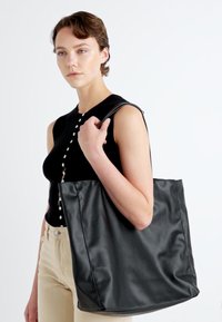 Črna usnjena torba z gladko teksturo, širokim telesom in kratkimi ročaji. Model nosi brezrokavnik črn vrh in bež hlače.