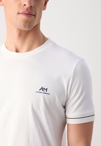T-shirt bianca in cotone con scollo tondo, caratterizzata da un piccolo logo blu navy sul petto e da strisce di accento blu navy sulle maniche.