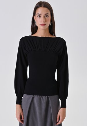 Donna che indossa un top aderente a maniche lunghe nero con dettagli arricciati e una gonna plissettata grigia, in piedi contro uno sfondo chiaro uniforme.