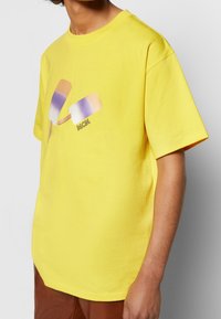 Camiseta de algodón amarilla con un gráfico de dos polos en tonos pastel, incluyendo rosa, blanco y púrpura, con el texto "MCM".