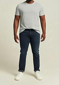 Homme portant un t-shirt à manches courtes gris clair, un jean foncé retroussé et des baskets blanches, debout devant un fond beige uni.