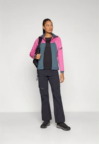 Dames outdoor jas in roze en teal, gecombineerd met een zwarte broek en laarzen. Voorzien van een capuchon, ritssluiting en functioneel ontwerp.