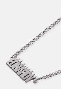 Collana in metallo argentato con un pendente piatto e a punta con la scritta "HARDY." Presenta una catena con texture e anelli intrecciati.