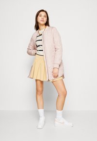 Veste matelassée rose clair avec un col rond, portée par-dessus un pull rayé et une jupe plissée jaune, assortie à des baskets blanches.
