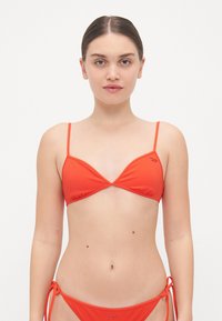 Rode bikini-top met driehoekige cups, minimale bandjes en geplooide details. Bevat een subtiele logo aan de linkerkant. Glad textuur.
