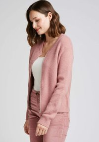 Cardigan tricoté rose clair avec fermeture éclair sur le devant, poignets et ourlet côtelés. Assortie avec un haut blanc et un pantalon en velours côtelé rose. Texture douce.