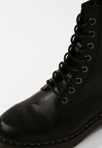 Zwarte leren veterschoen met metalen oogjes, ronde neus en platte zool met kleine metalen studs langs de onderrand.