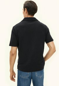 Czarna koszula z krótkim rękawem o fakturowanym wzorze, kołnierzykiem i luźnym krojem. W połączeniu z niebieskimi jeansami, widok z tyłu stroju.