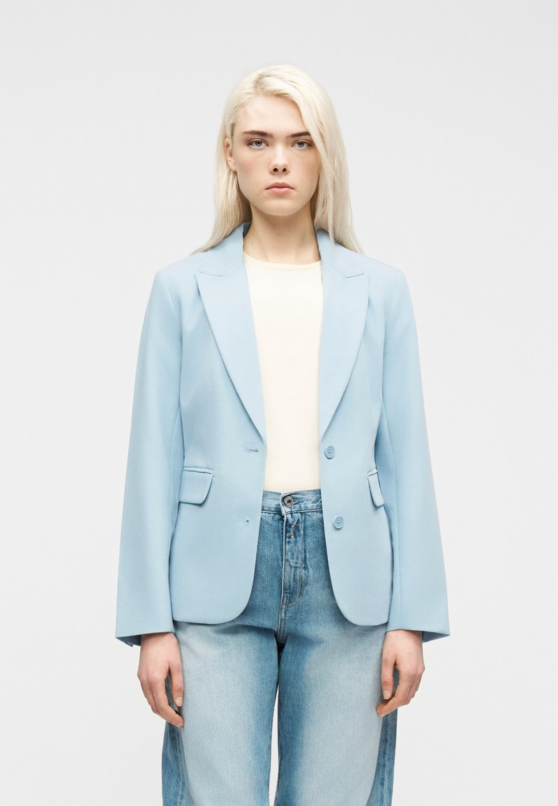 Νεαρή γυναίκα με μακριά ξανθά μαλλιά φοράει ανοιχτό μπλε σακάκι, λευκό τοπ και ανοιχτό μπλε τζιν, στέκεται μπροστά σε απλό φόντο.