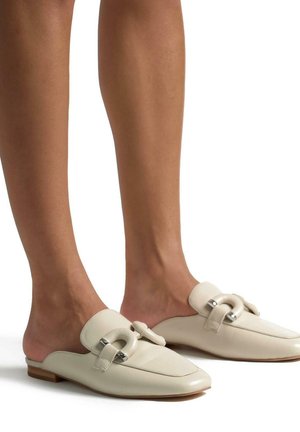 Mules en cuir beige avec un talon plat, un bout pointu et un détail en boucle argentée proéminent sur le cou-de-pied. Texture lisse et design minimaliste.