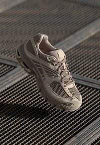 Beige tenisice s mrežicom i sintetičkim materijalom, zakrivljenog dizajna, s podstavljenim ovratnikom i teksturiranom potplatom. Imaju kontrastne bijele detalje i vezice.