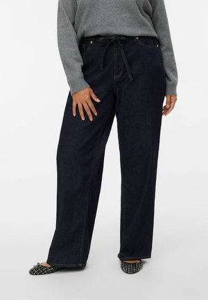 Personne portant un jean large bleu foncé avec une ceinture à nouer à la taille, un pull gris et des chaussures plates noires à motif, debout devant un fond uni.